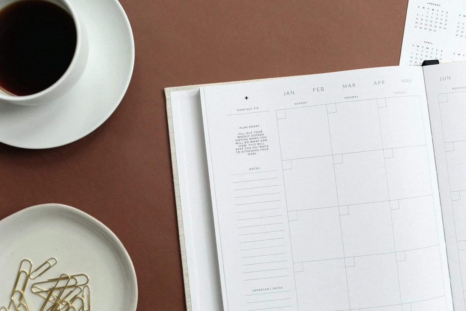 Open planner with monthly calendar spread, coffee cup, and gold paperclips on brown desk