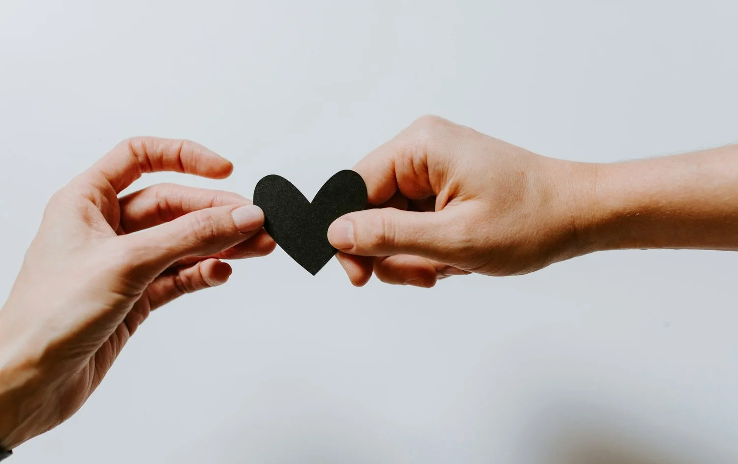 Two hands sharing a black paper heart against a white background