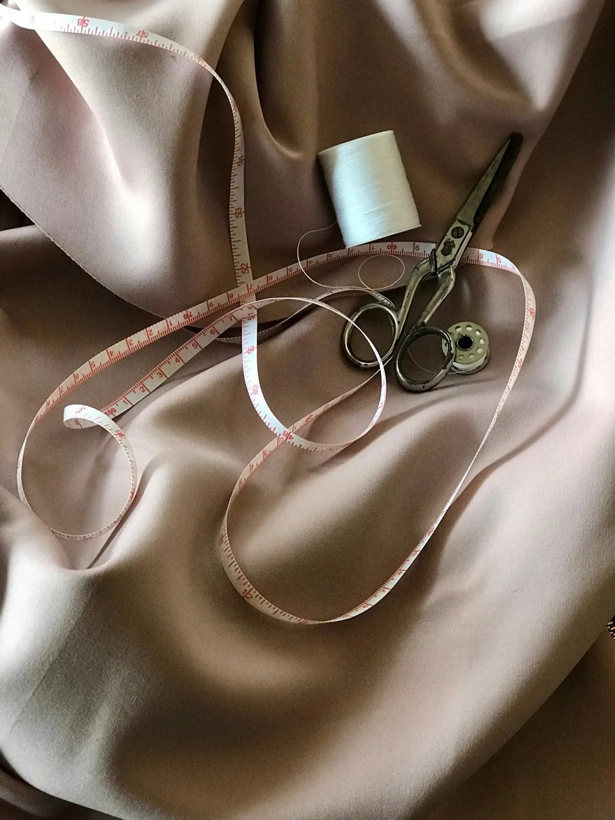 Sewing tools arranged on beige fabric including scissors, measuring tape, thread spool, and needle